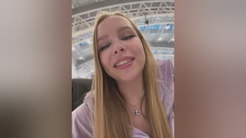 Media: Video of a smiling young Caucasian woman with long blonde hair, wearing a lavender shirt, taken indoors near a large, arched ceiling with blue accents.