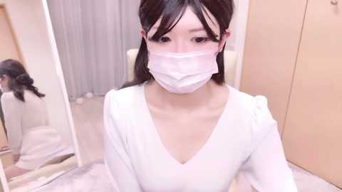 Media: Video of an Asian woman with straight black hair, wearing a white face mask and white top, sitting on a beige chair in a well-lit room with light-colored walls and a large mirror.