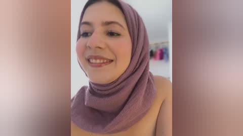 Media: Video of a smiling, fair-skinned woman with dark hair partially covered by a mauve hijab. She's indoors, wearing a light-colored top, with blurred clothing racks in the background.