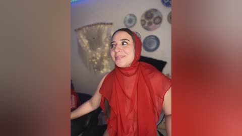 Media: Video of a smiling woman in a red hijab and blouse, sitting in a dimly lit room with a beige wall adorned with circular decorative items.