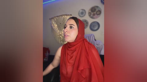 Media: A video of a young woman with light skin, wearing a red hijab and black top, standing in a dimly lit room with blue lights, framed art, and a gold fan on the wall.