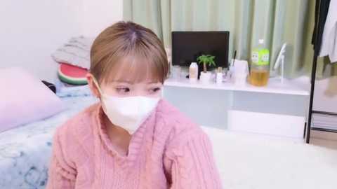 Media: Video of a young East Asian girl with short brown hair, wearing a pink sweater and white face mask, sitting in a pastel-colored bedroom with a bed, desk, and green curtains.