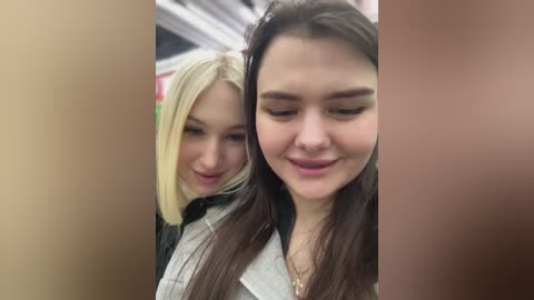 Media: Video of two young women with light skin tones and long hair; one blonde, one brunette. They have casual, friendly expressions and are standing close together, possibly in a shopping mall.