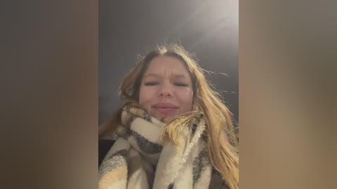 Media: A video of a fair-skinned woman with long, blonde hair, wearing a fur-trimmed coat, her eyes closed, looking serene, standing in a dimly lit room.