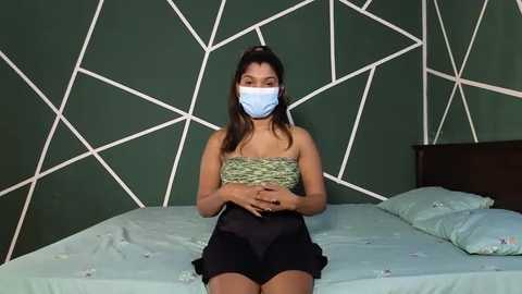 Media: Video of a South Asian woman with medium brown skin, wearing a blue mask, green and beige patterned bandeau top, black shorts, sitting on a bed with teal bedding, against a green wall with white geometric patterns.