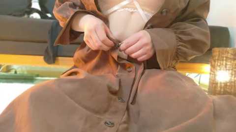 Media: Video of a woman with light skin, seated in a brown robe, being helped by another person with darker skin. Background shows a modern, indoor setting with a wicker lamp.