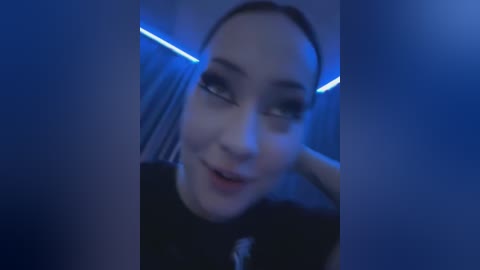 Media: Video of a young woman with fair skin, dark hair, and blue eyeshadow, wearing a black top, captured in a dimly lit room with blue lighting, giving a moody, mysterious vibe.