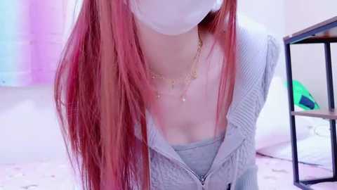 Media: Video of a young woman with long, straight, pink hair and a white face mask, wearing a light gray ribbed sweater with a zipper and a gold chain necklace. Background features a pastel-colored room with a black metal shelf and a green object.