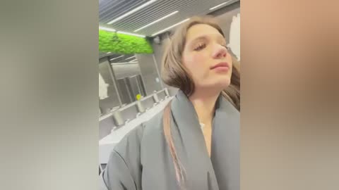 Media: Video of a woman with long brown hair, wearing a gray robe, standing in a modern bathroom with green wall accents, white sinks, and metallic fixtures.