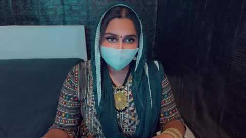 Media: Video of a woman in traditional attire, wearing a colorful, patterned shawl over a teal headscarf and a face mask, seated against a dark brick wall.