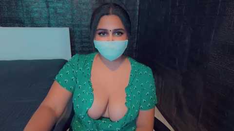 Media: Video of a plus-size woman with dark skin, large breasts, wearing a green floral dress with a deep neckline, blue face mask, and black hair tied back, sitting on a bed with dark walls.