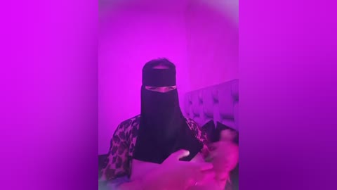 Media: A video featuring a person wearing a black mask with leopard print fabric, bathed in a purple light, standing in a dimly lit room with a bed and pillows in the background.