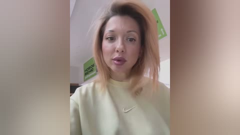 Media: Video of a woman with shoulder-length, light brown hair, wearing a beige sweatshirt, looking directly at the camera with a neutral expression. Background features a green poster and beige wall.