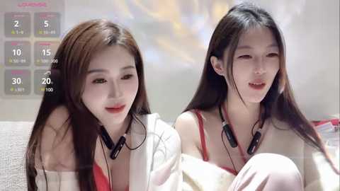 Media: Video of two young East Asian women with long black hair, wearing headphones, sitting on a couch in a bright, modern room with a calendar on the wall.