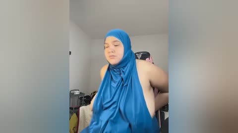 Media: Video of a young woman with fair skin and a slender build, wearing a bright blue hijab and a sleeveless top, standing in a minimalist, white-walled room with a cluttered desk in the background.