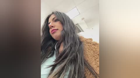 Media: Video of a woman with long, dark, wavy hair, wearing a light-colored top, and a brown jacket. She stands in a modern, white-walled room with fluorescent lighting.