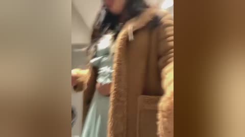 Media: A blurry video of a person with long dark hair wearing a light green dress and a brown fur coat, holding a drink, indoors with beige walls.