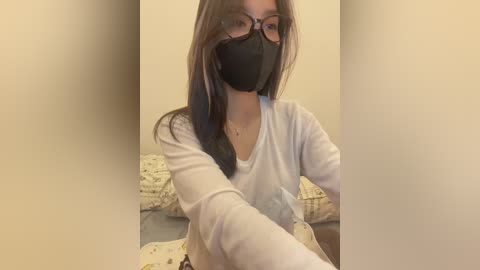 Media: A video of a young Asian woman with long, straight black hair and glasses, wearing a white long-sleeve shirt and black mask, seated on a bed with patterned bedding.