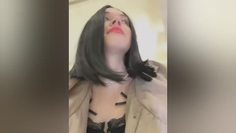 Media: Video of a young woman with shoulder-length black hair, fair skin, and red lipstick, wearing a beige jacket, black lace bra, and black tape across her chest. The background is blurred, suggesting an indoor setting.