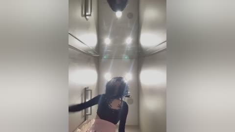 Media: A video of a person with shoulder-length curly hair, wearing a black top and pink skirt, captured from a low angle, looking up at a ceiling with multiple bright lights. The background is a modern, minimalist room with white walls and a wooden floor.