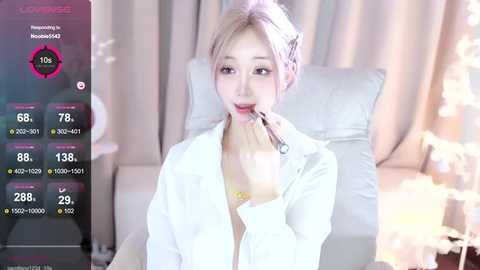 Media: A video of a young woman with light skin and platinum blonde hair, wearing a white blouse, applying makeup. She sits in a modern, softly lit room with beige curtains and a white couch.