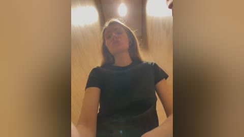 Media: Video of a young Caucasian woman with light brown hair, wearing a black t-shirt, sitting with legs apart in a dimly lit, wooden-paneled room, capturing a candid and intimate moment.