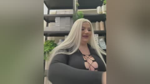 Media: Video of a plus-sized woman with long, platinum blonde hair, wearing a black, lace-up top, standing in a cluttered, indoor room with shelves and green plants in the background.