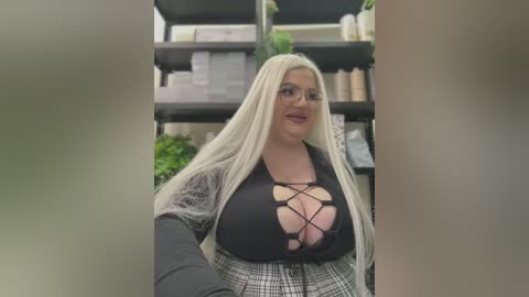 Media: Video of a plus-sized woman with long platinum blonde hair, wearing a black lace-up top revealing ample cleavage and a plaid skirt. She stands in a cluttered, dimly lit room with shelves filled with various items.