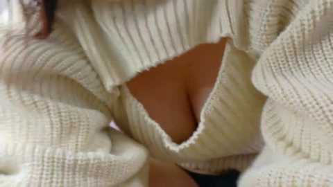 Media: Close-up video of a woman's upper body in a cream-colored knitted sweater with a large cutout revealing cleavage. The sweater is made of thick, textured yarn, and the woman's skin tone is light.