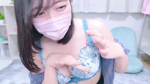 Media: Video of an Asian woman with long black hair, wearing a light pink face mask, blue floral bra, and a grey cardigan, adjusting her bra, in a room with white walls and a light blue cushion.