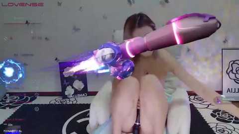 Media: Video of a young Asian woman with straight, shoulder-length brown hair, topless, wearing a pink, futuristic, high-tech gun on her head in a bedroom with a black and white floral rug.