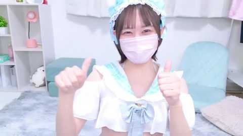 Media: Video of an Asian woman in a cute sailor outfit with a light blue headband and face mask, giving thumbs-up, standing in a bright, tidy room with pastel furniture.