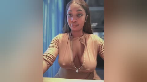 Media: Video of a dark-skinned woman with long, straight hair, wearing a tight, beige, long-sleeved dress with a deep V-neck, sitting at a table. Background shows blurred blue curtains and a bookshelf.