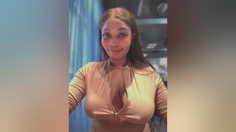 Media: Video of a dark-skinned woman with long straight hair, wearing a tight, peach-colored, zip-up top, revealing ample cleavage, standing indoors with a blue curtain and bookshelf background.