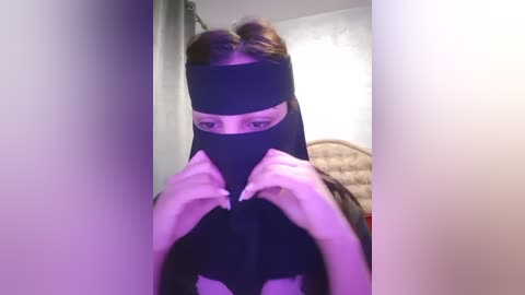 Media: A video of a woman with light skin and dark hair, wearing a black mask, adjusting it with her hands, against a gray background with a faint purple hue.