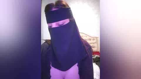 Media: Video of a person wearing a black niqab with a slit for the eyes, standing in a dimly lit bedroom with a bed and a white wall.