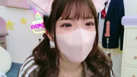 Media: Video of an East Asian woman with long brown hair in pigtails, wearing a pink mask, striped shirt, in a colorful, girly room with toys, a stuffed animal, and a neon sign.