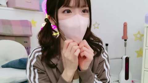 Media: A video of an Asian woman with long black hair in pigtails, wearing a white mask and a grey jacket with white stripes, in a brightly lit room with pastel furniture and a star-patterned wall.