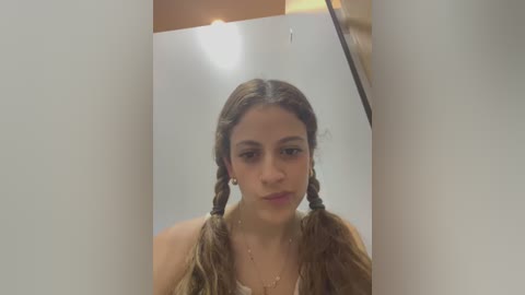 Media: A video of a young woman with light brown skin, long wavy hair in two braids, and a neutral expression, wearing a white top. The background is a white room with a partially visible wooden door.