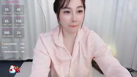 Media: A video of an East Asian woman with fair skin, dark hair in a bun, wearing a pink blouse, seated indoors, with a live stream overlay displaying views and comments.