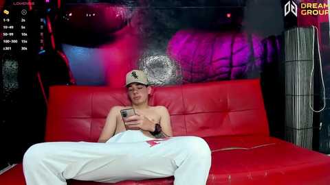 Media: A video of a young, shirtless man with a silver hat, white pants, and red couch in a dimly lit room with purple and red lights.