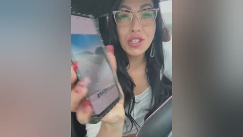 Media: Video of a young woman with long black hair, glasses, and hoop earrings, holding a smartphone mirror selfie, partially topless, showing medium-sized breasts.