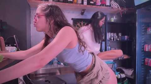 Media: Video of two young women in a kitchen; one with glasses and a blue tank top, the other in a white top, both bending over a counter.