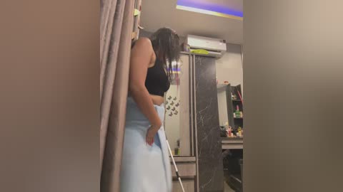 Media: Video of a woman with medium skin tone, medium build, wearing a black tank top and light blue skirt, leaning on a wall, in a modern kitchen with dark cabinets, white appliances, and a colorful mural.