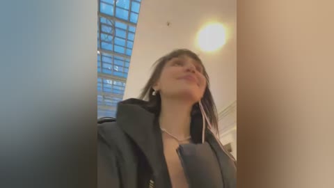 Media: A video of a woman with straight brown hair and a black coat, looking upward, in a modern, well-lit room with a glass block wall and a bright ceiling light.