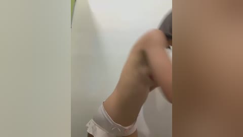 Media: A blurry video of a woman with a slender physique, wearing white shorts and a black headband, bending over in a corner, possibly in a bathroom with a pale green wall and a green plant.