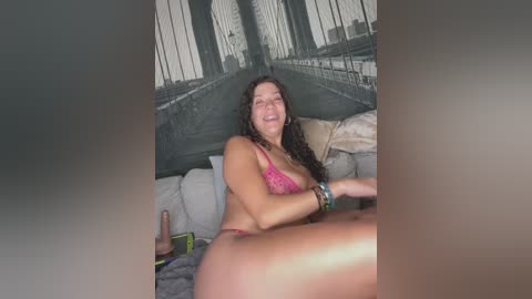 Media: Video of a smiling, light-skinned woman with curly hair in a pink lace bra and panties, lying on a bed with a gray duvet. The background features a mural of the Brooklyn Bridge.