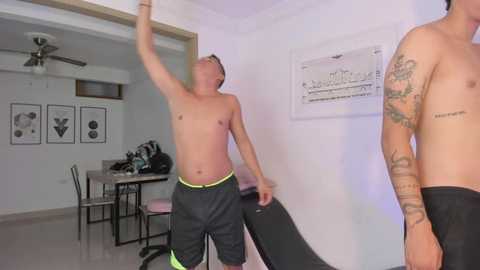 Media: Video of shirtless man with short hair and tattoos, wearing black shorts, dancing energetically in a modern, white-walled room with minimalist decor, including a ceiling fan, table, and framed art.