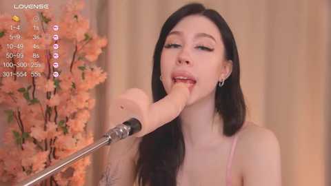 Media: A video of a young woman with long black hair and pale skin, wearing a pink spaghetti-strap top, seductively sucking on a large, realistic silicone dildo. Pink cherry blossoms and a digital camera are visible in the background.