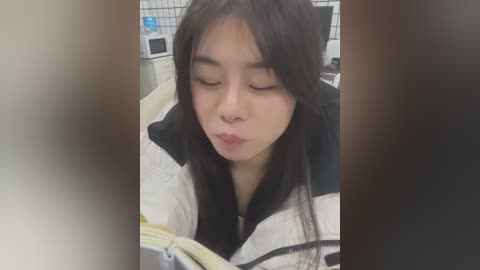 Media: Video of an Asian woman with long black hair, wearing a striped jacket, reading a book with eyes closed, in a kitchen with white tiled walls and a microwave.